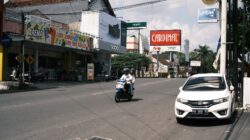 Jalanan ramai di Asia Tenggara dengan sepeda motor dan sebuah mobil yang terparkir di dekat toko-toko dan papan reklame.