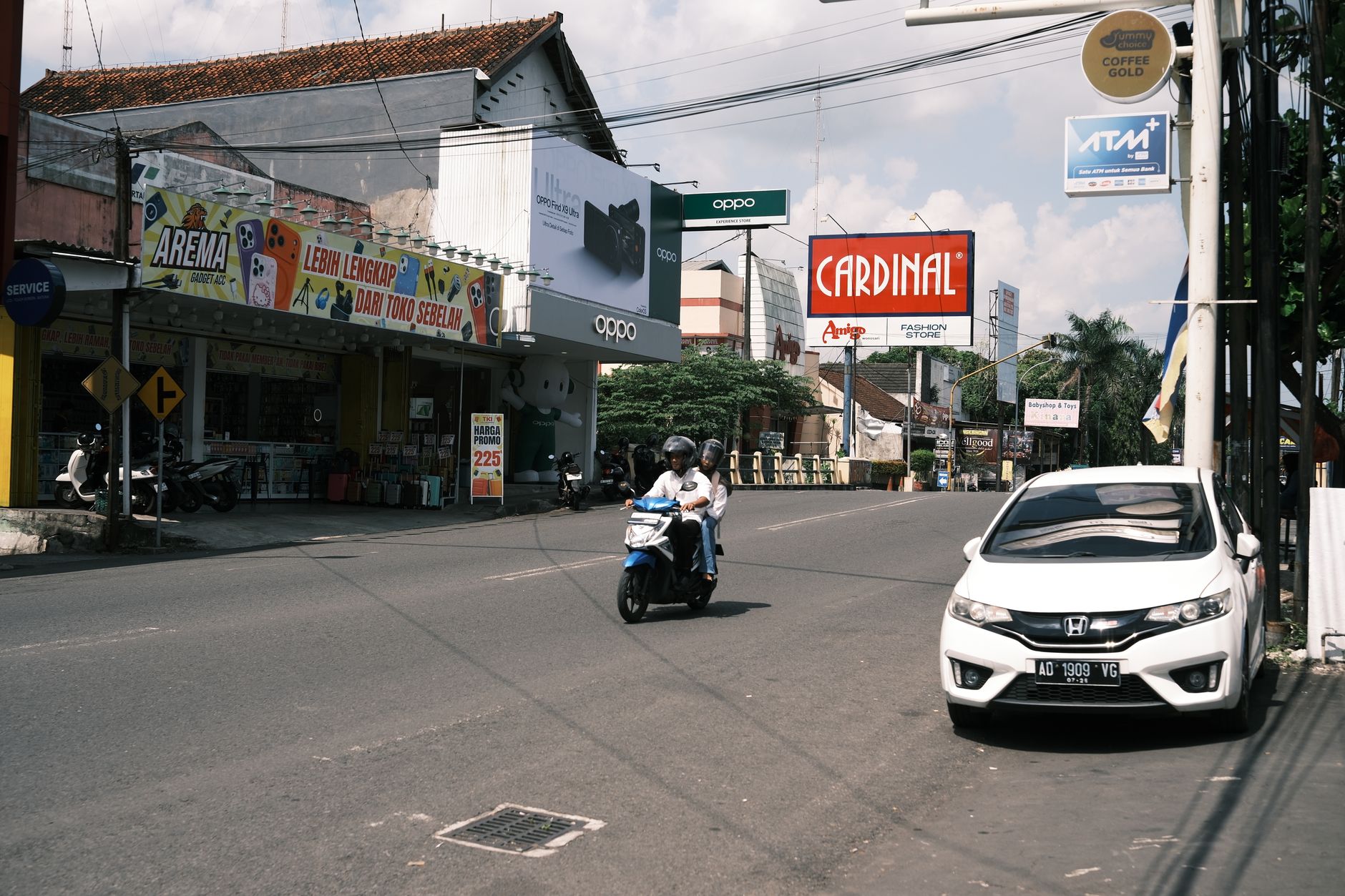 Jalanan ramai di Asia Tenggara dengan sepeda motor dan sebuah mobil yang terparkir di dekat toko-toko dan papan reklame.
