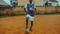Seorang pemain sepak bola remaja dalam seragam berpose dengan bola di lapangan tanah.
