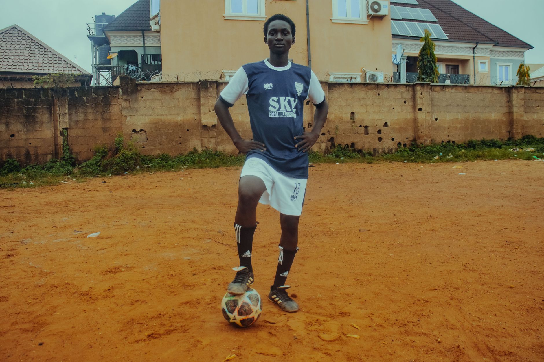Seorang pemain sepak bola remaja dalam seragam berpose dengan bola di lapangan tanah.