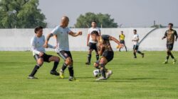 Pertandingan sepak bola dinamis dengan pemain bergerak di lapangan luar ruangan yang cerah.