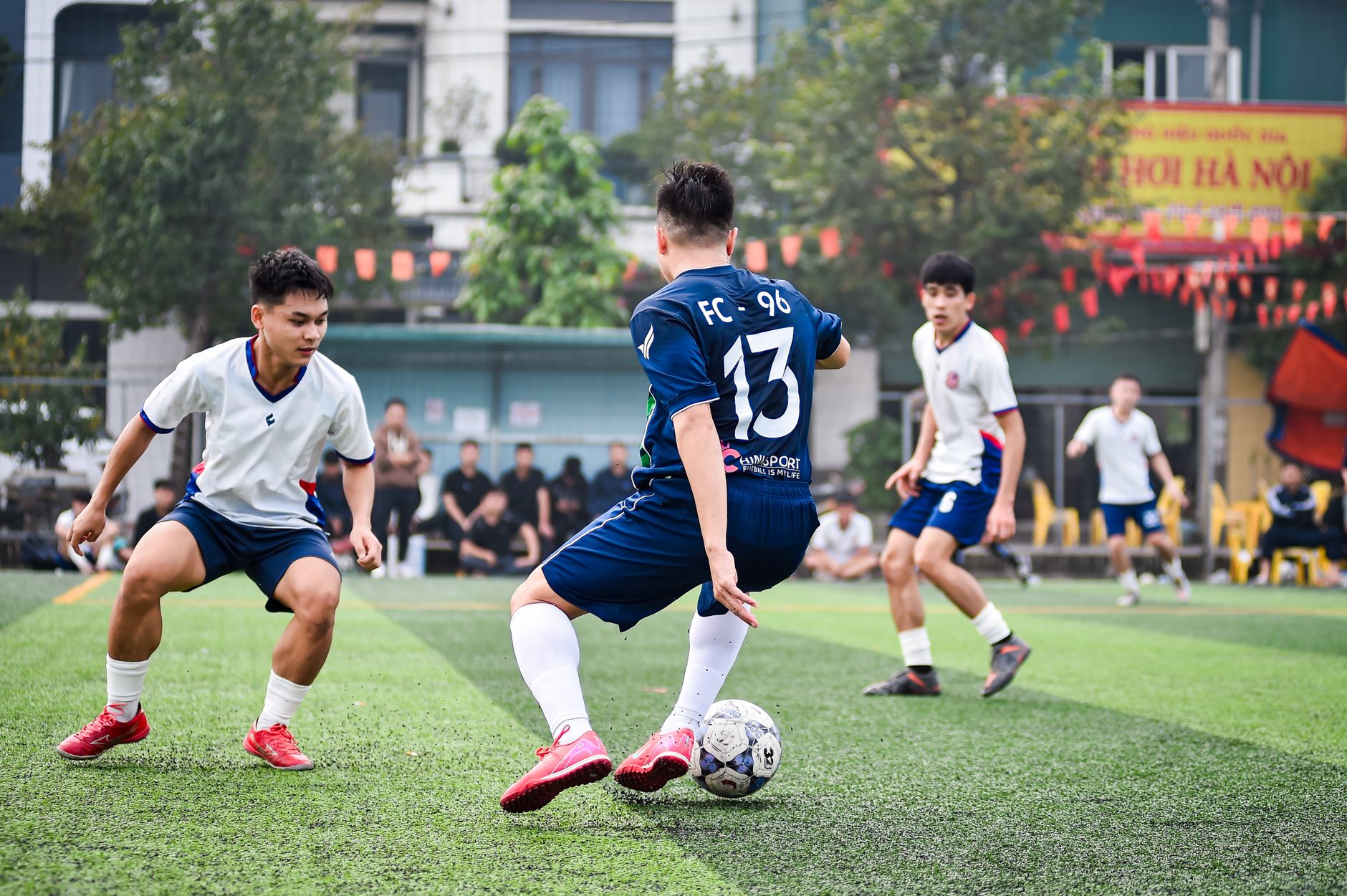 Pertandingan sepak bola dinamis di lapangan rumput buatan di Hanoi, yang menampilkan atletis dan semangat tim.
