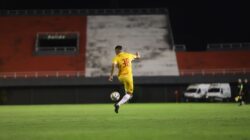 Pemain sepak bola dengan jersey kuning menendang bola di lapangan hijau saat pertandingan malam di bawah lampu.