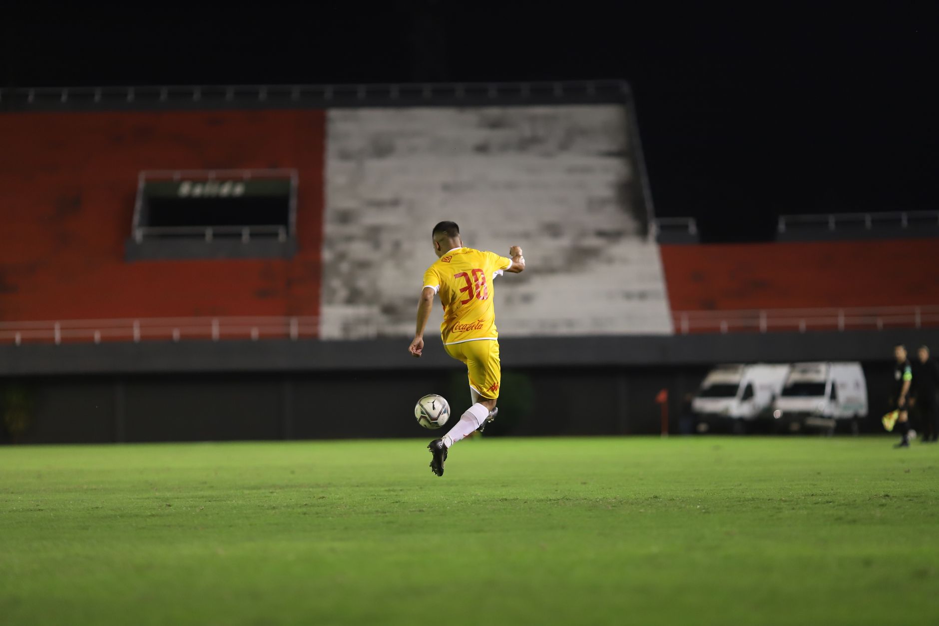 Pemain sepak bola dengan jersey kuning menendang bola di lapangan hijau saat pertandingan malam di bawah lampu.