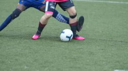 Dua pemain sepak bola bersaing memperebutkan bola di lapangan rumput selama pertandingan.