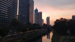 Pemandangan menakjubkan cakrawala Jakarta saat matahari terbenam, terpantul di kanal, menampilkan arsitektur modern.