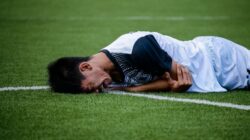 Pemain sepak bola tergeletak di lapangan kesakitan selama pertandingan, menunjukkan kemungkinan cedera.