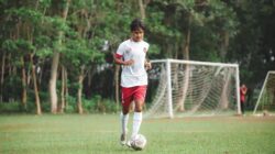 Pemain sepak bola remaja menggiring bola di lapangan sepak bola yang hijau subur.