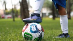 Seorang pemain sepak bola muda yang mengenakan kaus kaki putih berdiri di atas bola sepak warna-warni di luar ruangan.