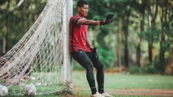 Seorang pemain sepak bola beristirahat di dekat tiang gawang di sebuah taman, mengenakan jersey merah.
