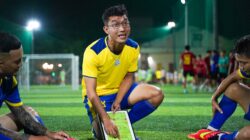 Seorang pelatih sepak bola merumuskan strategi di lapangan bersama pemain selama sesi permainan malam.