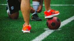 Close-up pemain sepak bola saat latihan, satu mengikat tali sepatu, yang lain menyeimbangkan bola di rumput.