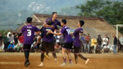 Pemain sepak bola merayakan gol dalam pertandingan luar ruangan yang berdebu dengan penonton.