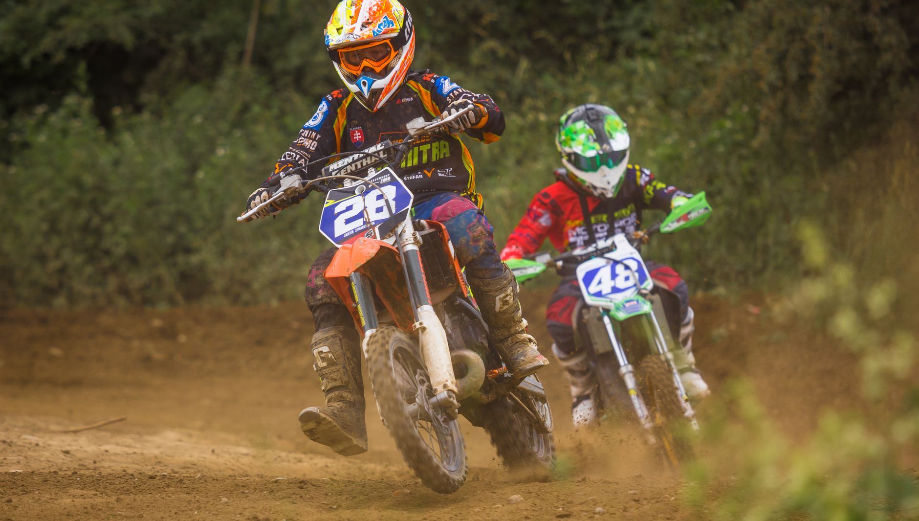Balapan motocross dinamis dengan dua pengendara yang melaju di jalur tanah mengenakan helm dan perlengkapan berwarna-warni.