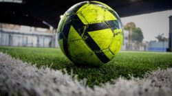 Gambar close-up bola sepak neon di lapangan rumput buatan di dalam ruangan.