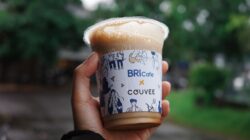 Close-up tangan memegang cangkir kopi es bermerek di luar ruangan, dengan latar belakang hijau buram yang cerah.