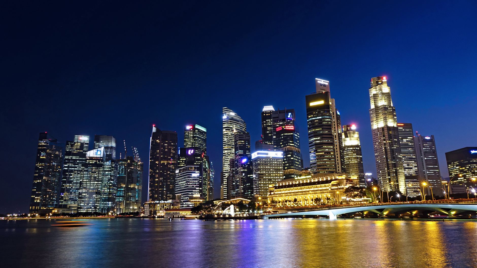 Pemandangan malam yang cerah dari cakrawala kota Singapura dengan gedung pencakar langit yang diterangi yang terpantul di air.