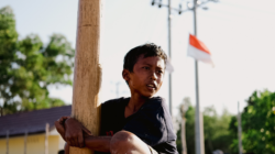 Seorang anak memanjat tiang kayu di lingkungan luar ruangan dengan bendera Indonesia terlihat di latar belakang.