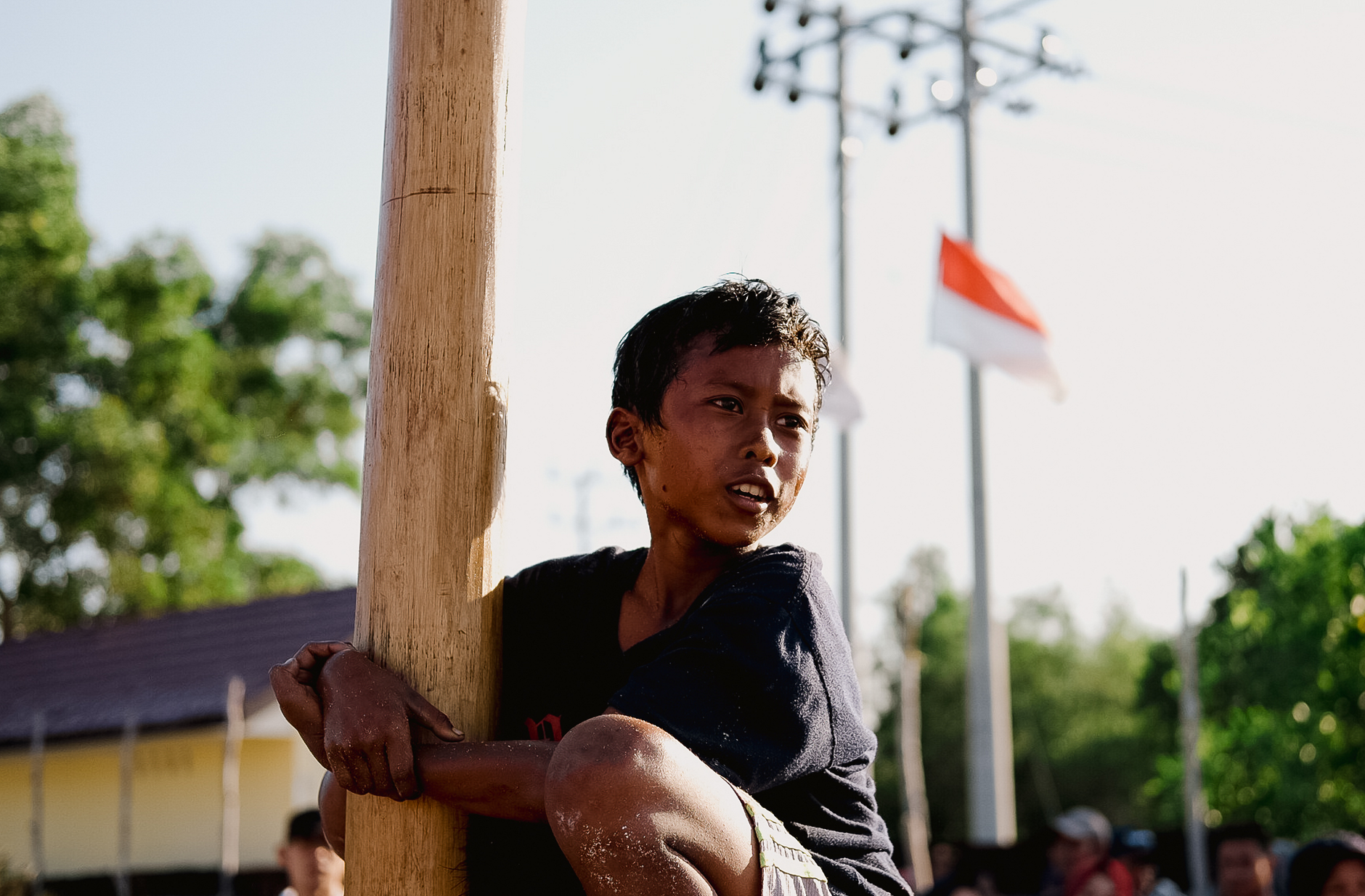 Seorang anak memanjat tiang kayu di lingkungan luar ruangan dengan bendera Indonesia terlihat di latar belakang.
