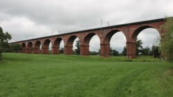 Jembatan kereta api bersejarah di Inggris yang membentang di atas ladang hijau subur di bawah langit berawan.