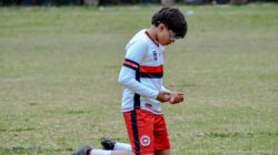 Seorang pemain sepak bola muda berpakaian seragam berlutut di lapangan rumput di luar ruangan.