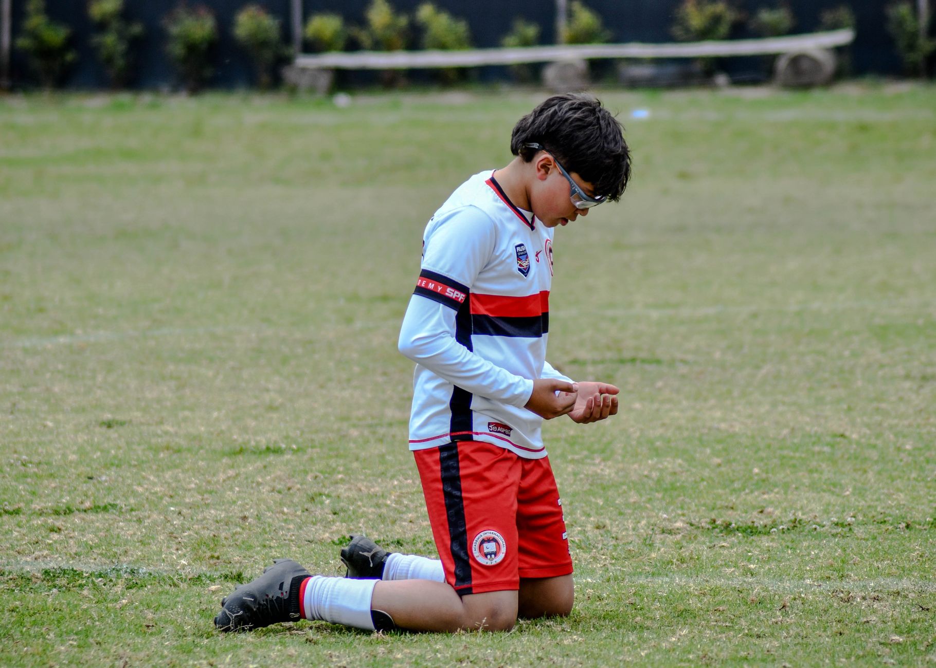 Seorang pemain sepak bola muda berpakaian seragam berlutut di lapangan rumput di luar ruangan.