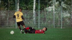 Dua pemain sepak bola dalam aksi dengan tekel dramatis di lapangan rumput selama pertandingan.