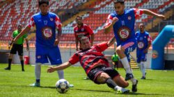 Pertandingan sepak bola seru dengan pemain yang bergerak di bawah sinar matahari yang cerah. Menangkap kerja sama tim dan semangat olahraga di lapangan.