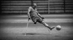 Tangkapan dinamis dari pemain sepak bola adaptif yang menendang bola dengan kruk di dalam ruangan dalam hitam dan putih.