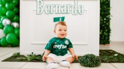 Bayi yang bahagia duduk dan tersenyum dalam jersey hijau di acara dalam ruangan yang didekorasi.