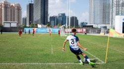Seorang pemain sepak bola muda mengambil tendangan sudut pada hari yang cerah di Dubai dengan gedung pencakar langit di latar belakang.