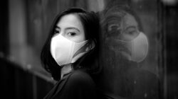 Potret hitam putih yang mencolok dari seorang wanita yang mengenakan masker wajah, merefleksikan pada sebuah jendela.