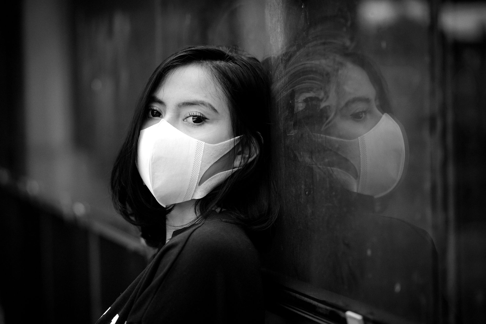 Potret hitam putih yang mencolok dari seorang wanita yang mengenakan masker wajah, merefleksikan pada sebuah jendela.