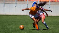Dua pemain sepak bola wanita bersaing sengit untuk merebut bola selama pertandingan di luar ruangan yang cerah.