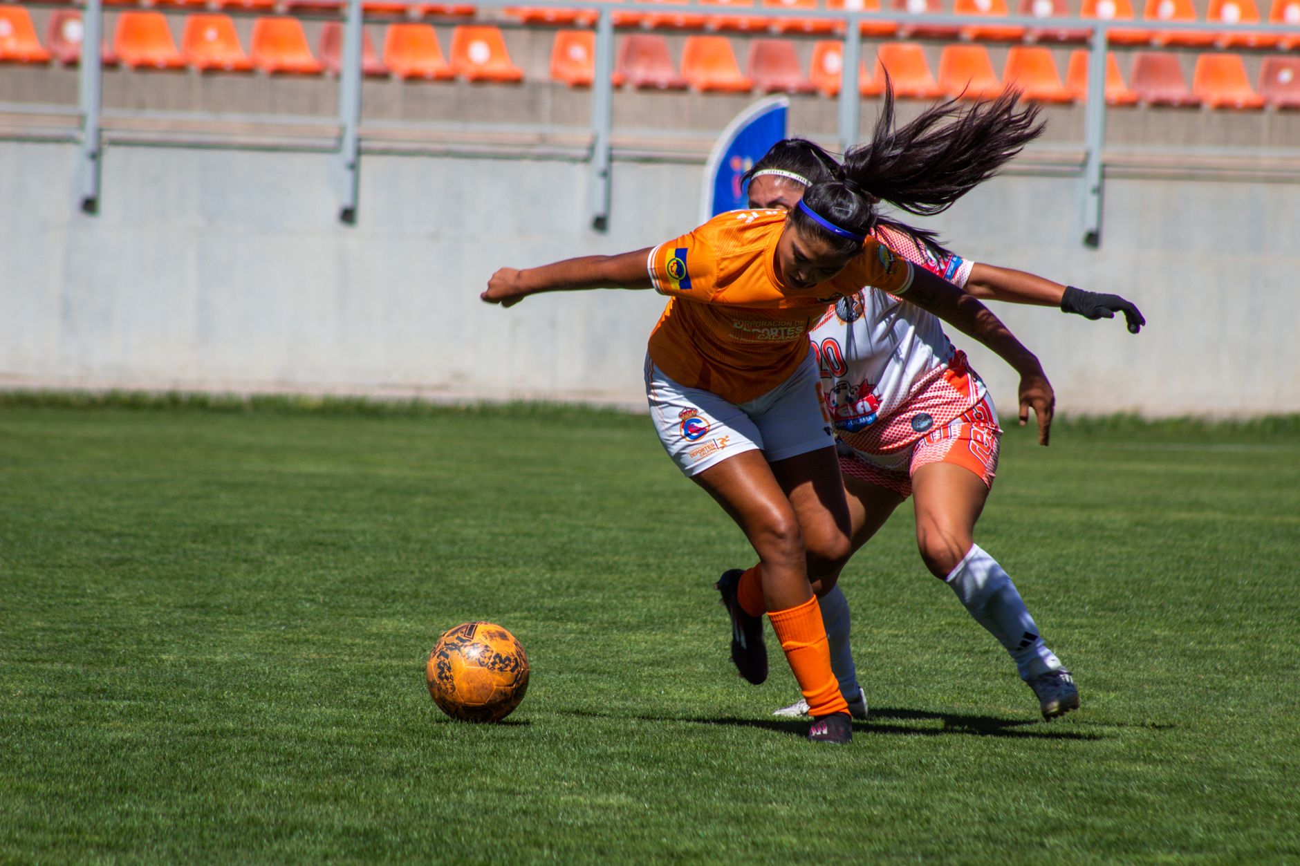 Dua pemain sepak bola wanita bersaing sengit untuk merebut bola selama pertandingan di luar ruangan yang cerah.