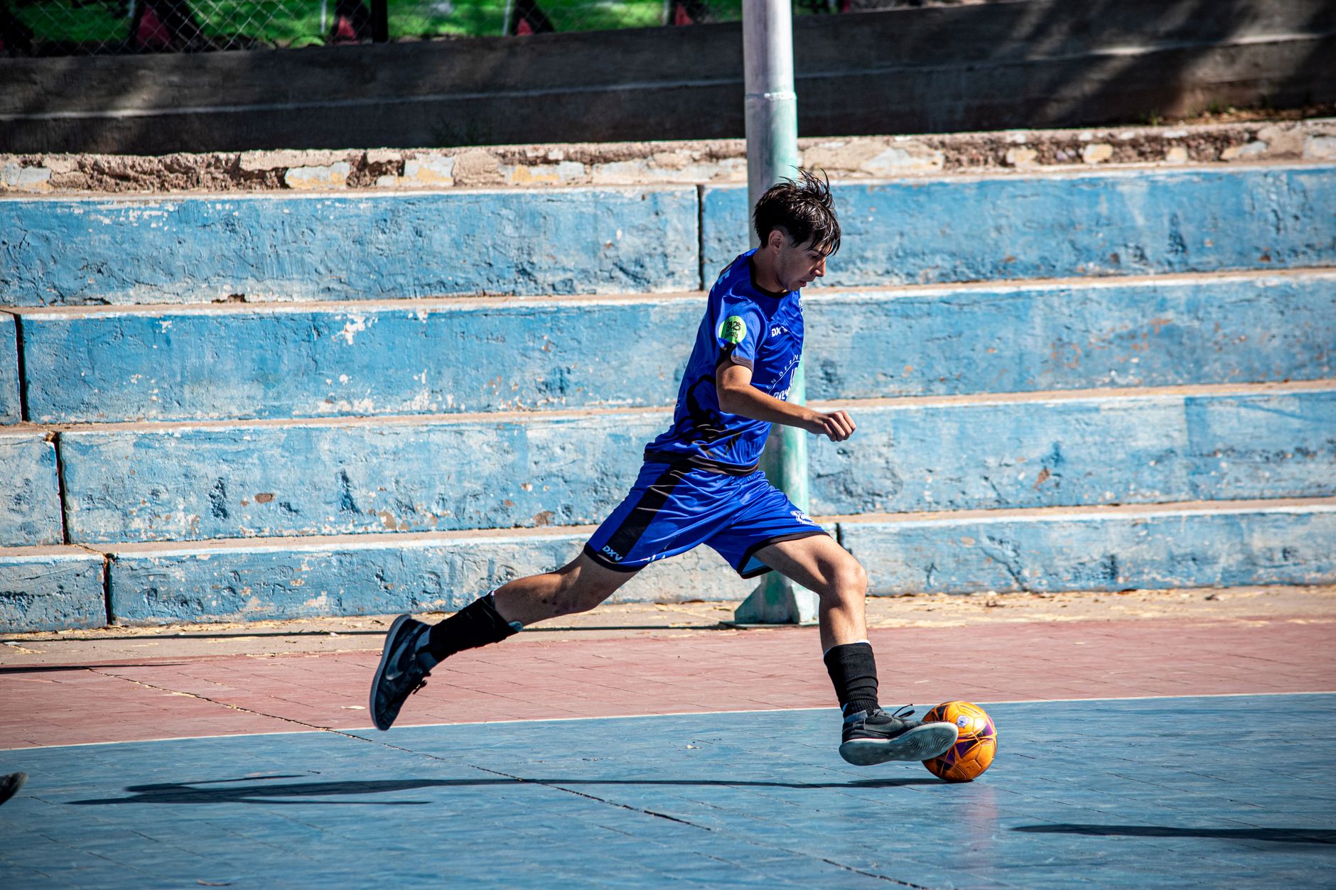 Bocah remaja dengan jersey biru bermain sepak bola di lapangan outdoor, sedang dalam aksi.