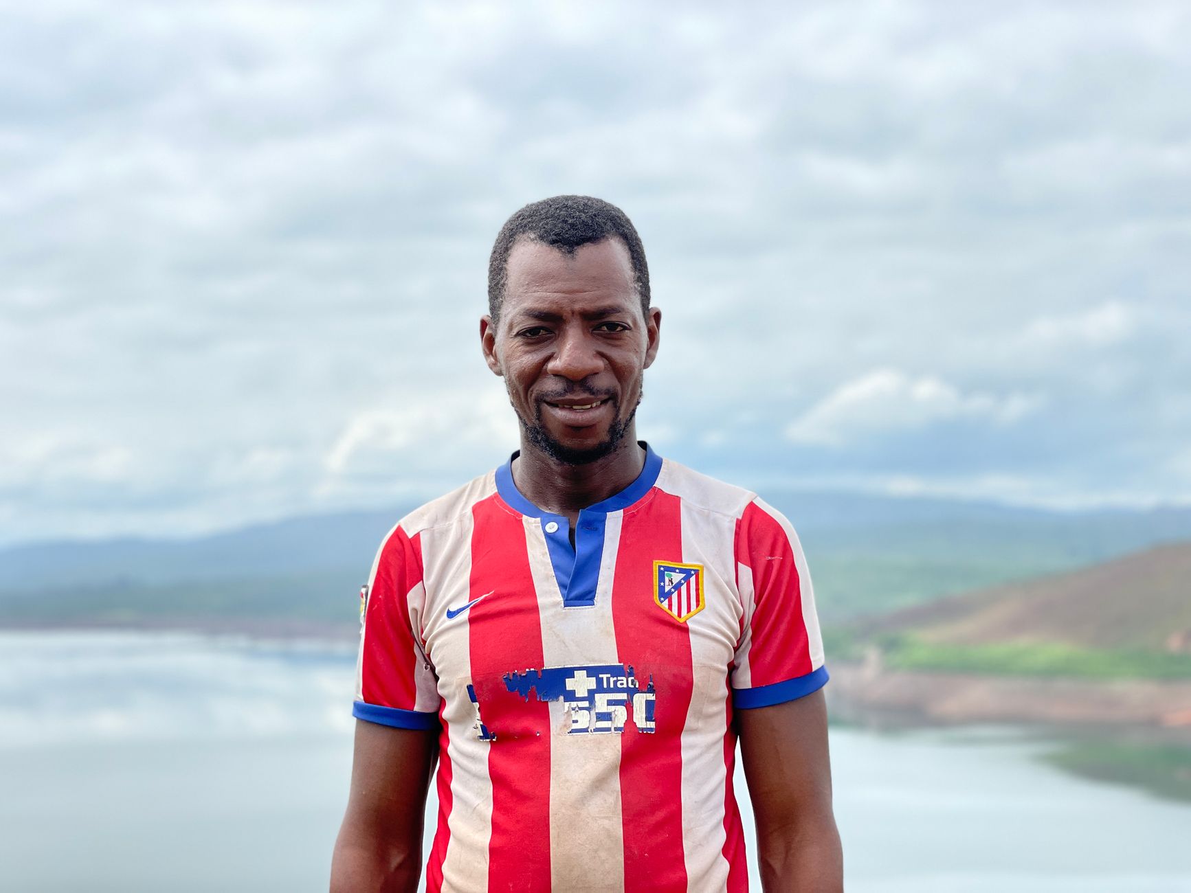 Potret seorang pria Afrika yang tersenyum dengan jersey bergaris di latar belakang pemandangan alam di Guinea.