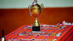 Piala emas dengan pita dan medali di atas meja kotak, melambangkan kemenangan dan pencapaian.