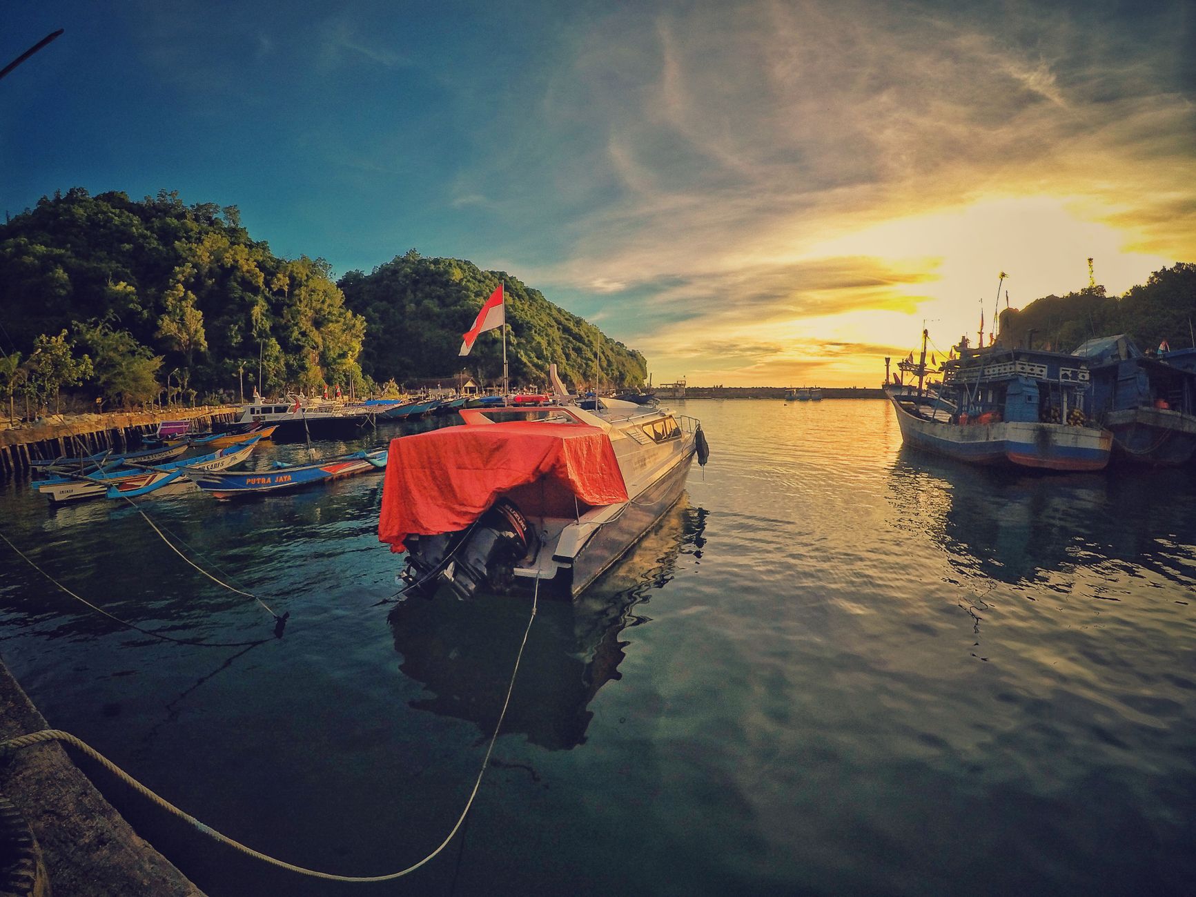 Pemandangan matahari terbenam yang menakjubkan di Pelabuhan Girisubo, Indonesia, dengan perahu dan bukit hijau yang subur.