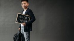 Seorang bocah muda dengan ransel memegang papan tulis bertuliskan 'Halo Sekolah', siap untuk sekolah.