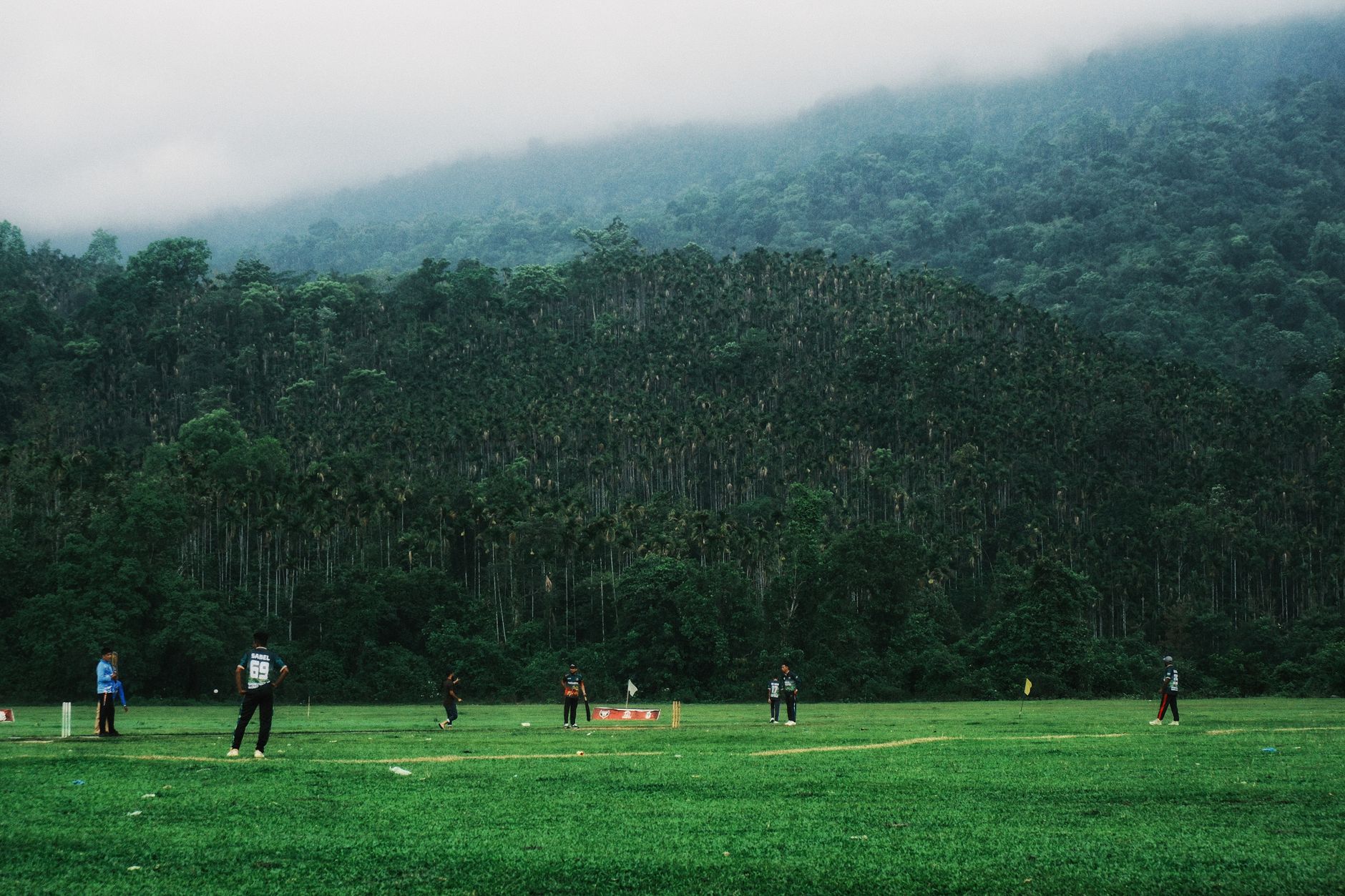 Pemain kriket bermain di lapangan hijau subur dikelilingi hutan di Sylhet, Bangladesh.