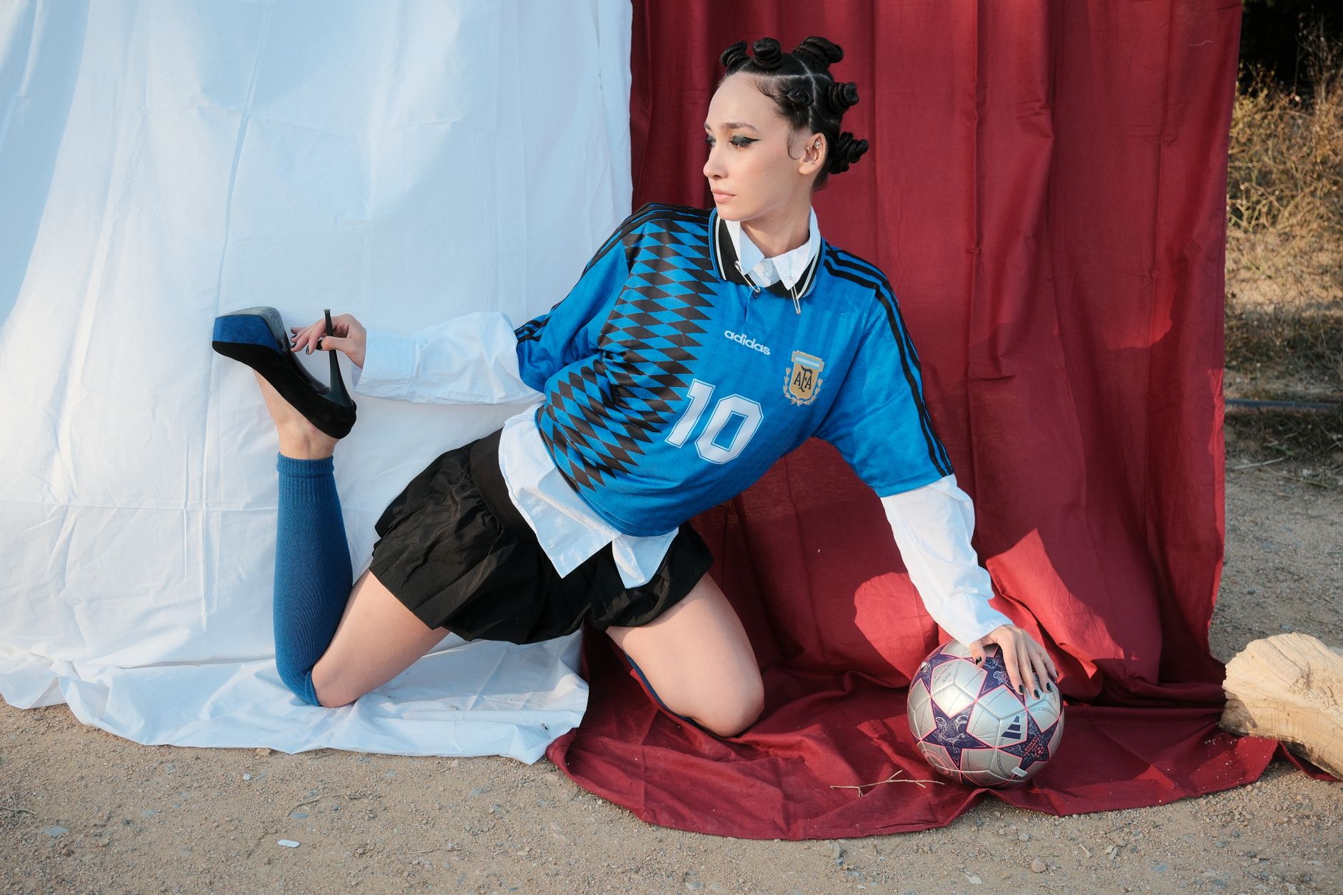 Model stylish berpose dengan bola sepak dan sepatu hak tinggi, memadukan olahraga dan mode dalam suasana luar ruangan yang unik.