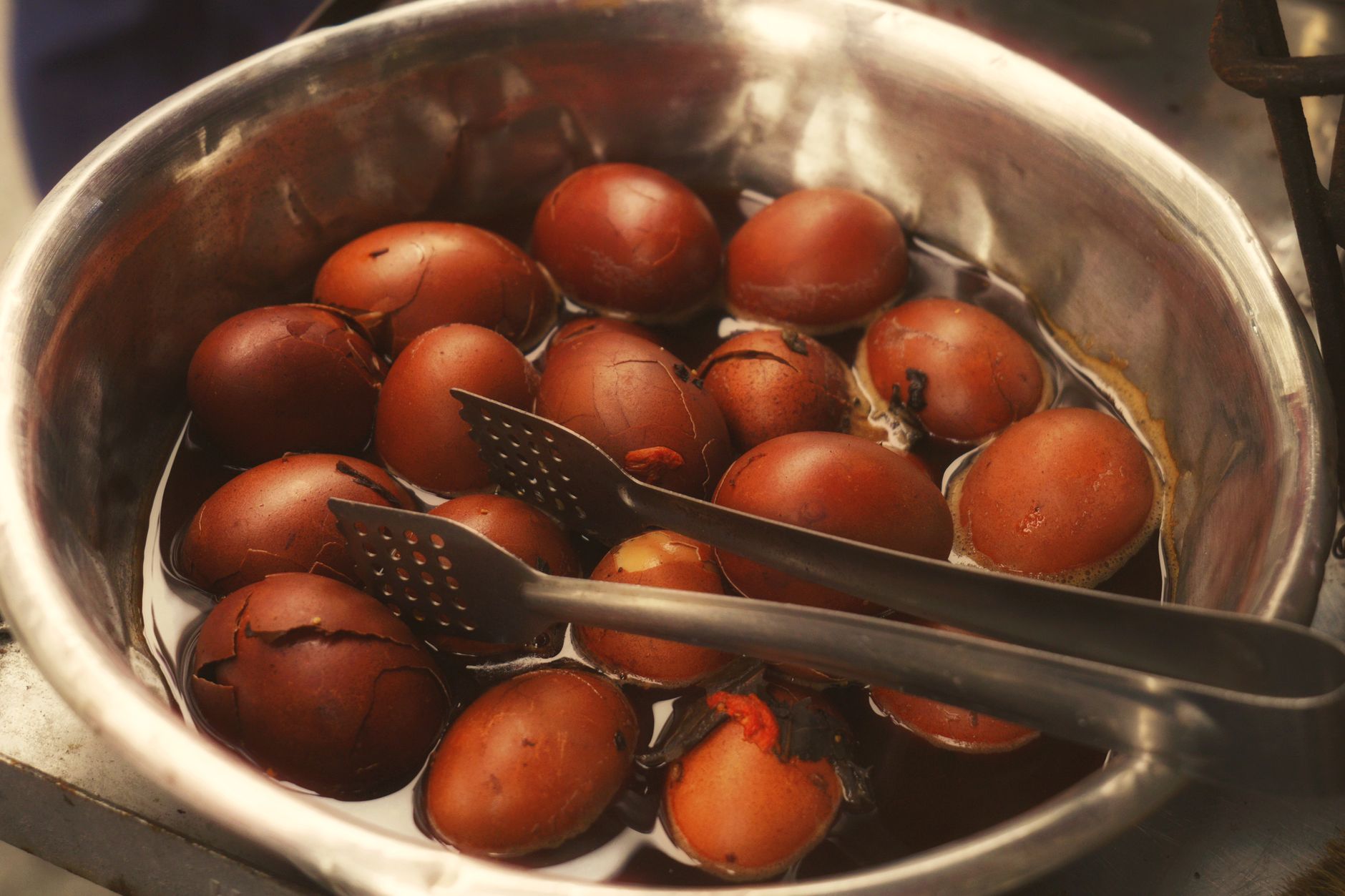 Telur rebus segar dalam rendaman teh tradisional dengan penjepit, menyoroti budaya kuliner Asia.
