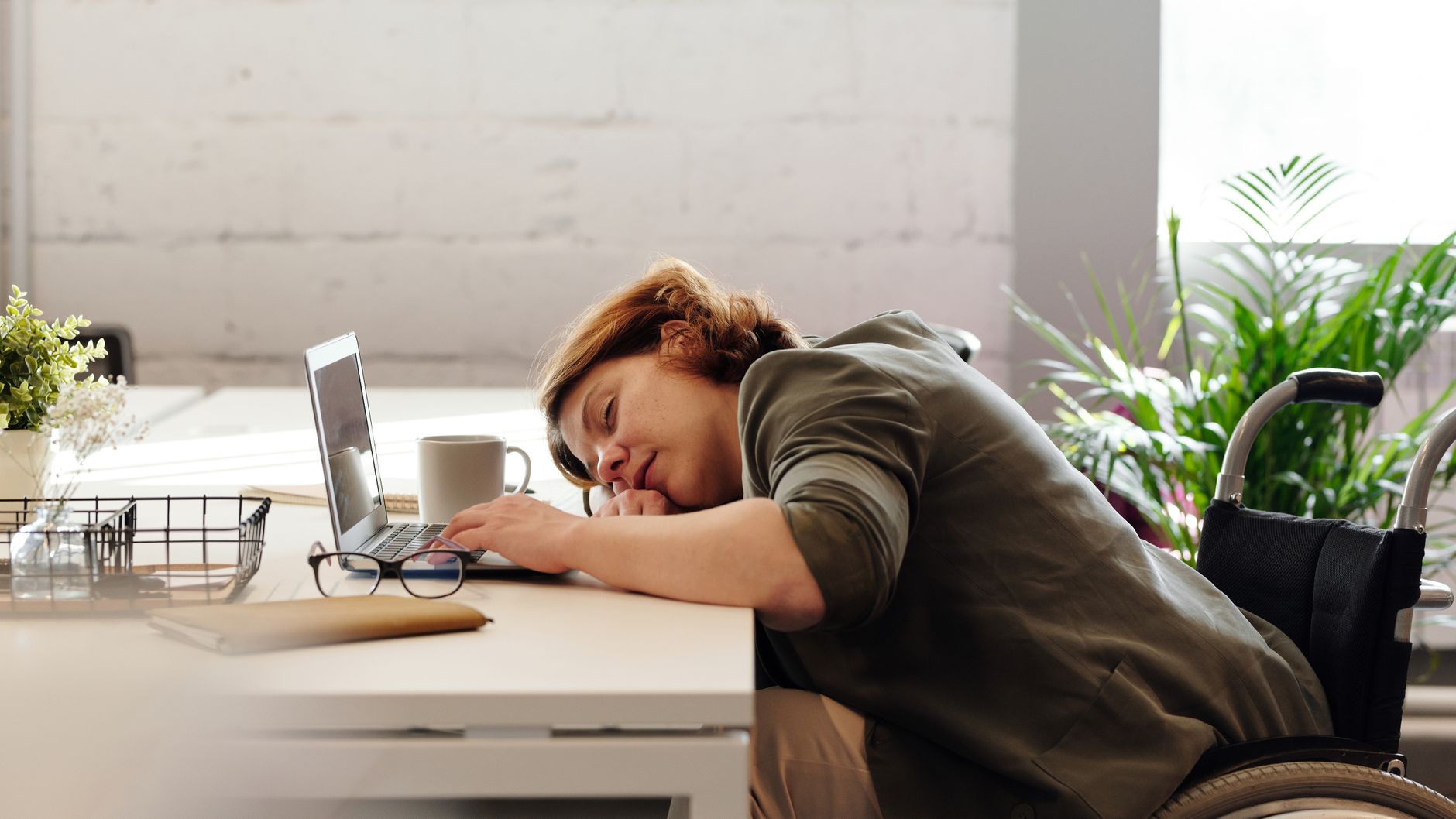 Wanita di kursi roda yang tertidur di meja dengan laptop, menggambarkan kelelahan dan tantangan bekerja jarak jauh.