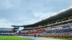 Sebuah stadion penuh dengan penonton antusias menyaksikan acara olahraga langsung di bawah cahaya yang terang.