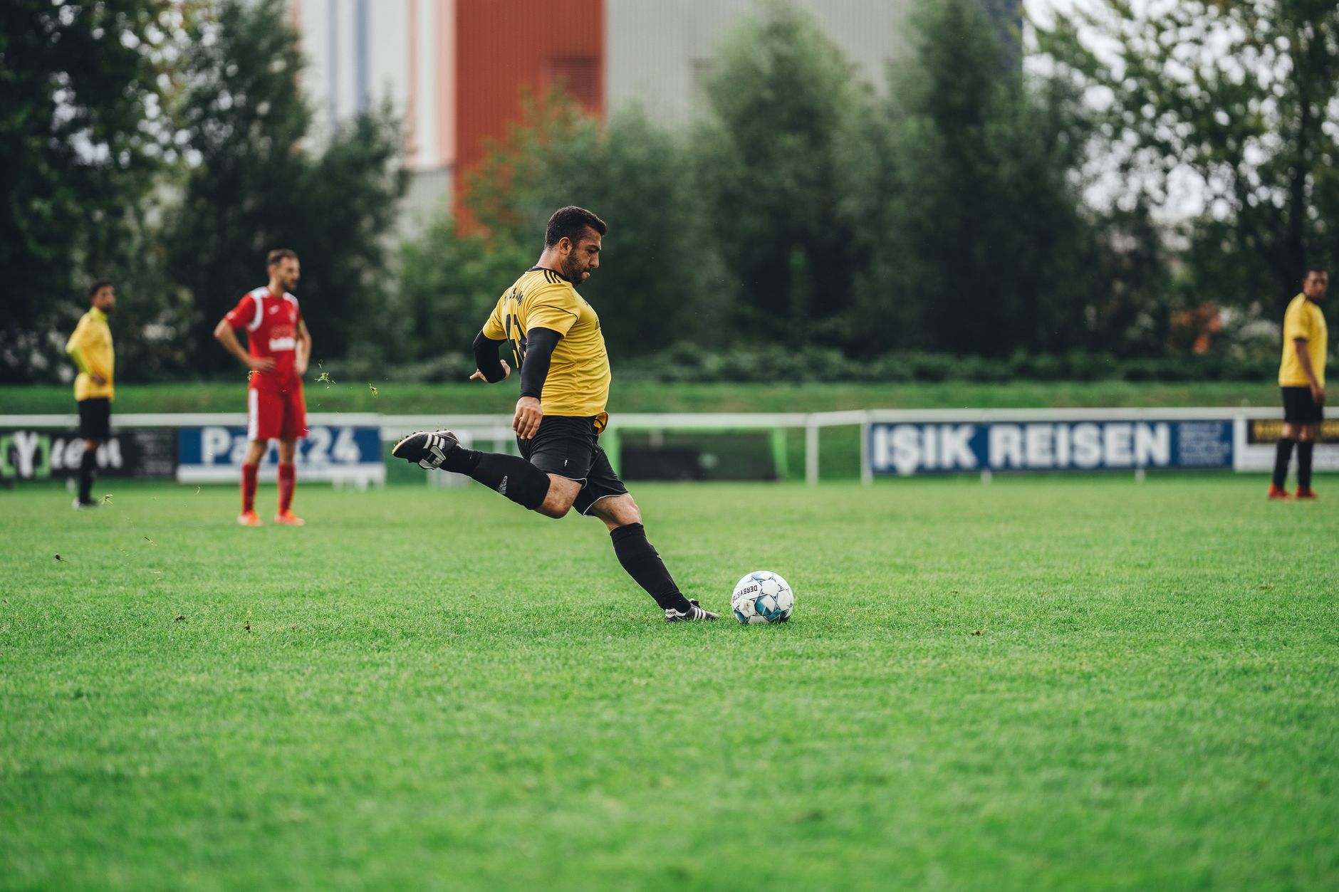 Seorang pemain sepak bola dalam seragam kuning menendang bola selama pertandingan di lapangan hijau luar.
