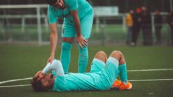 Seorang pemain sepak bola berbaring di lapangan rumput yang dip安kan oleh rekan satu tim dalam pakaian olahraga.