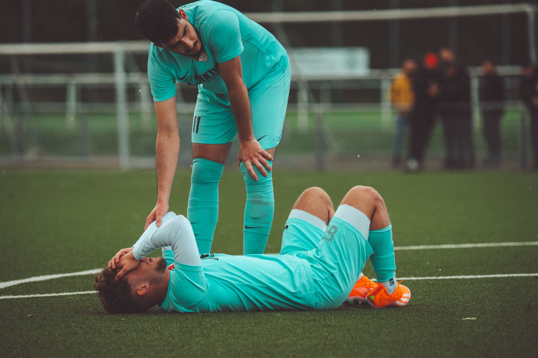 Seorang pemain sepak bola berbaring di lapangan rumput yang dip安kan oleh rekan satu tim dalam pakaian olahraga.