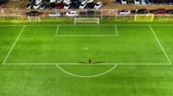 Pemandangan udara lapangan sepak bola di malam hari di Jakarta dengan seorang pemain sendirian yang melemparkan bayangan di bawah lampu stadion.