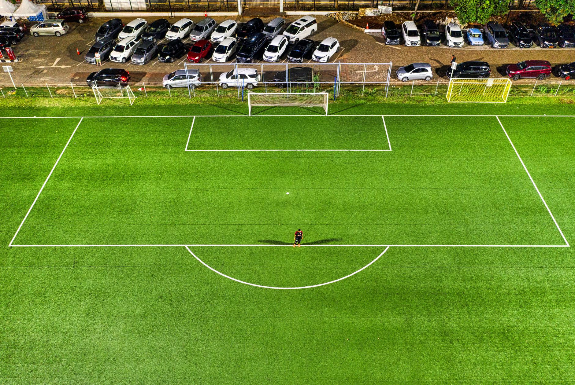 Pemandangan udara lapangan sepak bola di malam hari di Jakarta dengan seorang pemain sendirian yang melemparkan bayangan di bawah lampu stadion.